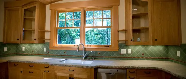 Wood kitchen cabinets around a double window with a green tile backsplash and open corner shelving